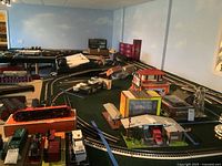 Wide view of green platform with complex 3-rail track layout including various curves, sidings, and structures including a gas station and warehouse.