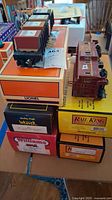 Photo showing a stack of model train car boxes from Lionel, Rail King, Weaver, and Williams with two visible model train cars: a caboose and a boxcar on top of their boxes.