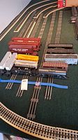 Photo showing 13 model train cars on a green felt display with railroad tracks beneath, multiple boxcars, a tanker, a gondola visible. Tracks not included.