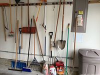Photo showing multiple yard and garden tools hanging on wall and standing against it including shovels, rakes, garden fork, brooms, red gas can and containers at base