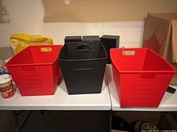 Three plastic storage bins displayed on table: two red and one black. All have handles and appear stackable.