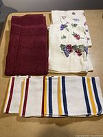 Photo showing burgundy hand towels, white hand towels with grape cluster embroidery, and striped dish cloths spread out on a surface.