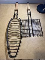 Two BBQ grill baskets side by side on a flat surface, one fish-shaped and one square grid shaped, both with wooden handles.