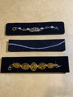 Three bracelets displayed on black velvet holders: amber-colored oval stone bracelet with toggle clasp at bottom, thin silver-tone multi-strand chain bracelet in middle, multi-stone opalescent and amethyst bracelet at top.