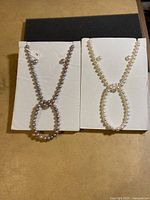 Two pearl bead necklace sets displayed on white card backing; one set with white pearls, the other with grayish pearls.