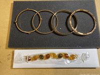 Four gold-tone bangles on foam pad and one multicolored stone-link bracelet sealed in plastic on white foam