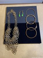 Photo of the full set: multi-chain gold-tone necklace, three gold-tone bangles, and green drop earrings on black velvet background.