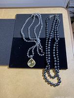 Two necklaces laid out on black and gray background. Clear bead necklace has yellow teardrop pendant. Black necklace has pearlescent beads.