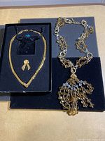 Two necklaces and a pin displayed on black velvet trays; one gold-tone chain necklace in V shape, one beaded necklace with amber and clear beads and cluster pendant, and a small gold-tone ribbon pin.