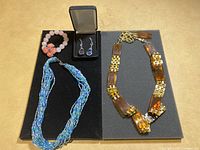 Overview photo showing all jewelry pieces on black display board: tortoiseshell-style necklace, blue beaded necklace, pink beaded bracelet with floral accent, and drop earrings in black box.