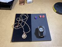 Men's watch, beaded necklace with teardrop pendant, purple ring, and red flower earrings displayed on black panels.