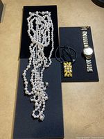 Pearl-style multi-strand necklaces, black cord necklace with enamel pendant, and silver-tone wristwatch displayed on black foam pads.