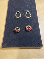 Two teardrop-shaped gold hoop earrings paired with two rings, one featuring cluster of light rhinestones, other with red rhinestones, on dark fabric.