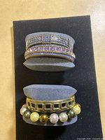 Four bracelets displayed on gray foam holders, showing varied designs and colors including purple, blue, pearl beads and gold-tone metal.