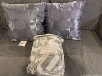 Two grey square pillows with a pattern of large circles and a packaged grey and white chevron patterned throw on a grey couch.