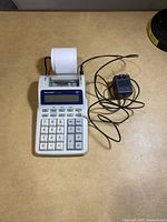 Full view of Sharp EL-1701C calculator with power adapter and paper roll on table.