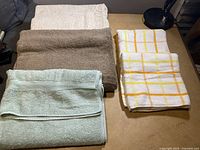 Five towels laid out on a flat surface; three solid colored bath towels in off-white, light brown, and light green-blue, alongside two white hand towels with yellow-orange plaid pattern.