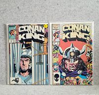Two Conan the King comic books, issue 97 and 37, displayed side by side in plastic sleeves on a gray carpet background.