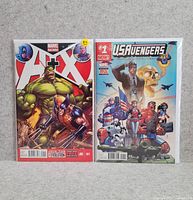 Two Marvel comic books displayed front-facing, showing covers of A+X #1 with Hulk and Wolverine and U.S.Avengers #1 with multiple superheroes and flag imagery, bagged upright on neutral background.