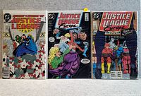 Three Justice League comic books laid on a flat surface, all in protective plastic covers. The comics show colorful front covers with detailed superhero illustrations.