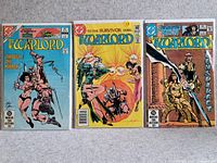 Photo of three Warlord comic books laid flat side-by-side on carpet. Covers visible showing titles: 'Through the Mirror,' 'To the Survivor Goes...,' and 'Conspiracy.'