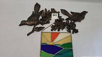 Photo shows two dark brown hard plastic bird wall hangings resting on branches with detailed flowers, chained to a stained glass sunset-themed sun catcher with hills and a blue lake/river design.
