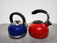 Photo showing blue and coral red enamel kettles side by side on a grey surface, front angle