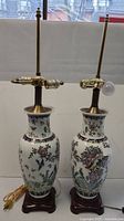 Front view of two tall ceramic lamps side by side showing painted floral and bird designs, wooden bases, brass tops, and electrical cords.