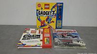 Three LEGO books with associated parts packaging placed on grey floor against white background. Gadgets book is boxed and unopened, Chain Reactions book with LEGO parts, and Contraptions book in resealed plastic packaging with gears and wheels visible.