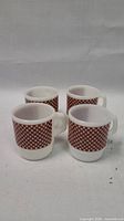 Front angled view of 4 white mugs with brown and white gingham checkerboard pattern around the body, showing handles and bases.