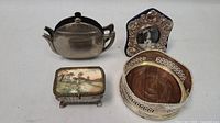 Photo showing all four vintage metal items: teapot napkin holder, glass and metal trinket box, photo frame with black and white photo, and metal wine bottle holder with wooden insert base.