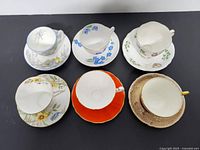 Six bone china tea cups with matching saucers in various floral and solid color designs laid out on black background.