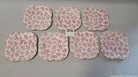 All seven small square china plates laid out showing red floral pattern and scalloped edges
