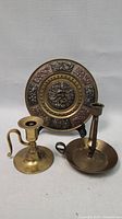 Three brass items: two candlestick holders and one decorative hanging plate on stand, shown together.