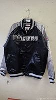 Front view of black satin Oakland Raiders jacket with silver sleeves and striped ribbing on collar, cuffs, and hem.