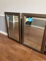 Two rectangular wall mirrors with brown frames resting against a wall on a wooden floor, showing front reflective surface.