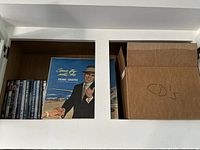 Wood cabinet shelf with DVDs in cases and a boxed vinyl album of Frank Sinatra's Come Fly With Me, along with a cardboard box labeled CDs