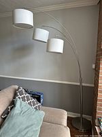 Full view of three-armed arched floor lamp positioned next to a couch, showing the metal base, three curved metal arms, and white fabric drum shades.