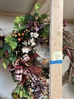 Close-up of a fall-themed wreath with artificial berries, leaves, small white flowers, and a plaid bow, hanging on a wooden rack.