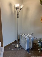 Two white Lasko box fans and a silver floor lamp in a corner of a room.