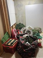 Wide view showing assorted Christmas decorations on the floor against a wall including wreaths, fabric bag with greenery, Christmas tree stand, and various bins.