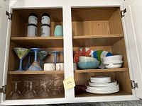 Kitchen cabinet open showing shelves with white reusable water bottles, colorful pottery martini glasses, Mikasa tea cups, Corelle plates and bowls, plastic and ceramic bowls and plates