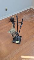 Metal fireplace tool set on wooden floor with power cords in background. Includes gloves hanging on the stand.