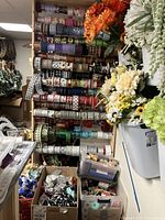 Wide view of wooden ribbon rack filled with multiple rolls of ribbons in various patterns and colors, plastic bins with additional ribbons and craft supplies below, and artificial flowers on the right side.