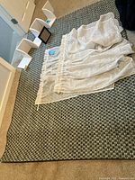 Overview of the rug, curtains laid atop it, white corner shelf pieces, and picture frames arranged on the floor.