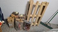 Overview of three bundles of firewood wrapped in plastic, bag of fire starter splints, small wooden pallet, and a green broom in background. Pen on floor for size reference.