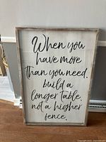 Front view of framed wall art with the saying 'When you have more than you need, build a longer table, not a higher fence.' The frame is wooden with a glass panel that is not attached.