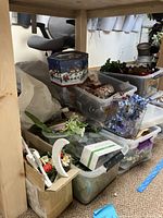 Wide view of plastic bins and boxes filled with wreath decorating supplies including artificial flowers and greenery under a wooden table.