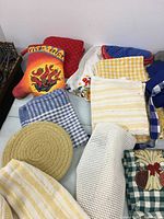 View of assorted kitchen towels, potholders, aprons and napkins in various colors and patterns - solid, checkered, striped, printed.