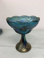 Side view of blue-green iridescent carnival glass pedestal candy dish showcasing grape and leaf relief pattern and scalloped rim.